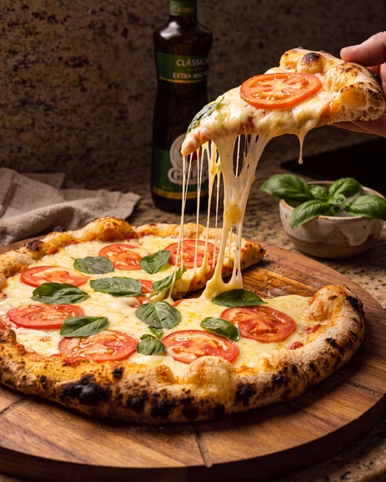 Fatia de pizza marguerita puxando o queijo derretido, com tomate e manjericão fresco, ao lado da pizza inteira na tábua